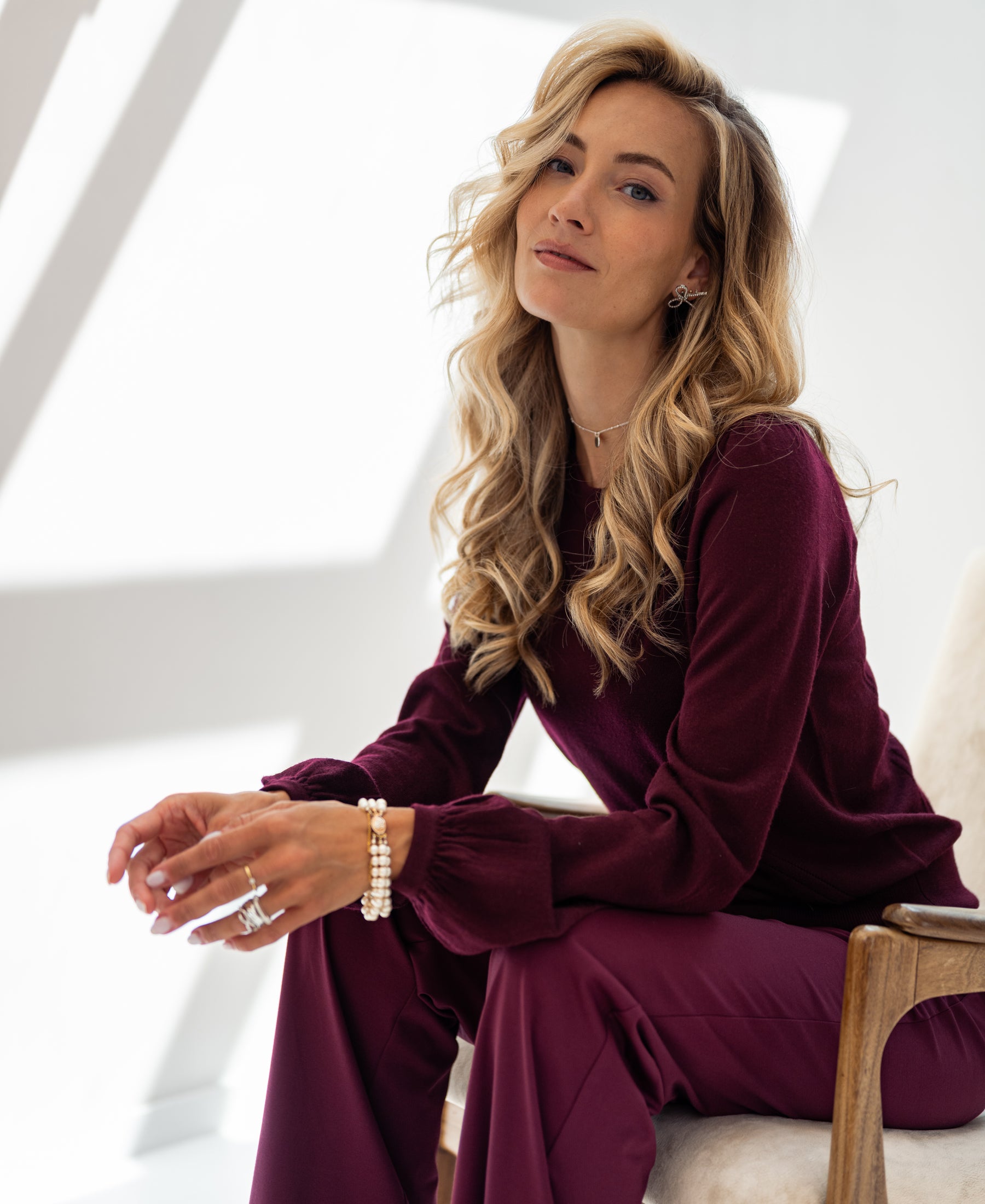 A woman wears a Bordeaux red LA COEUR sweater by PLEIN PUBLIQUE, shown seated and softly smiling in natural light.