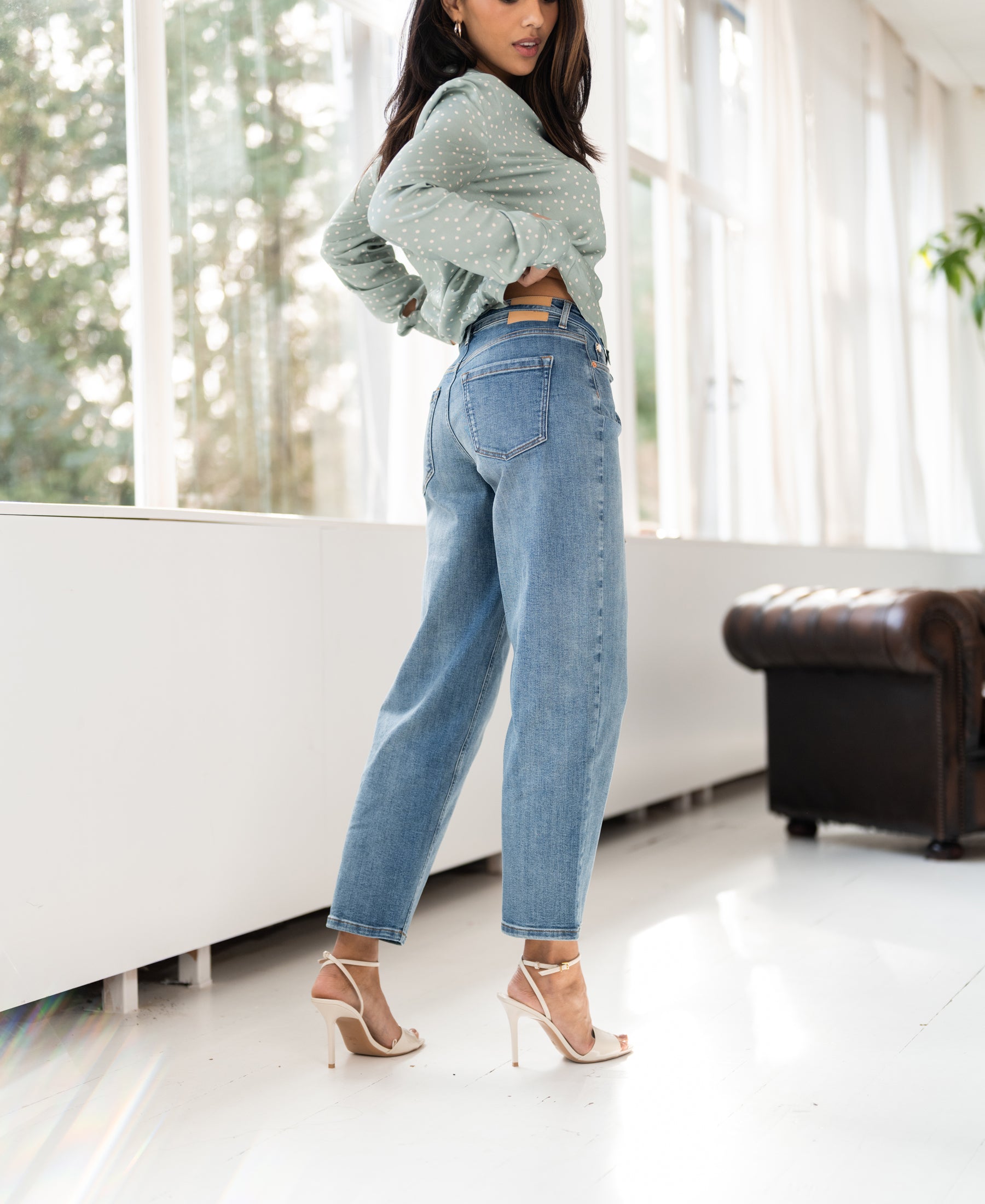 Woman wearing light green cropped jeans stands by large windows.