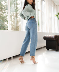 Woman wearing light green cropped jeans stands by large windows.