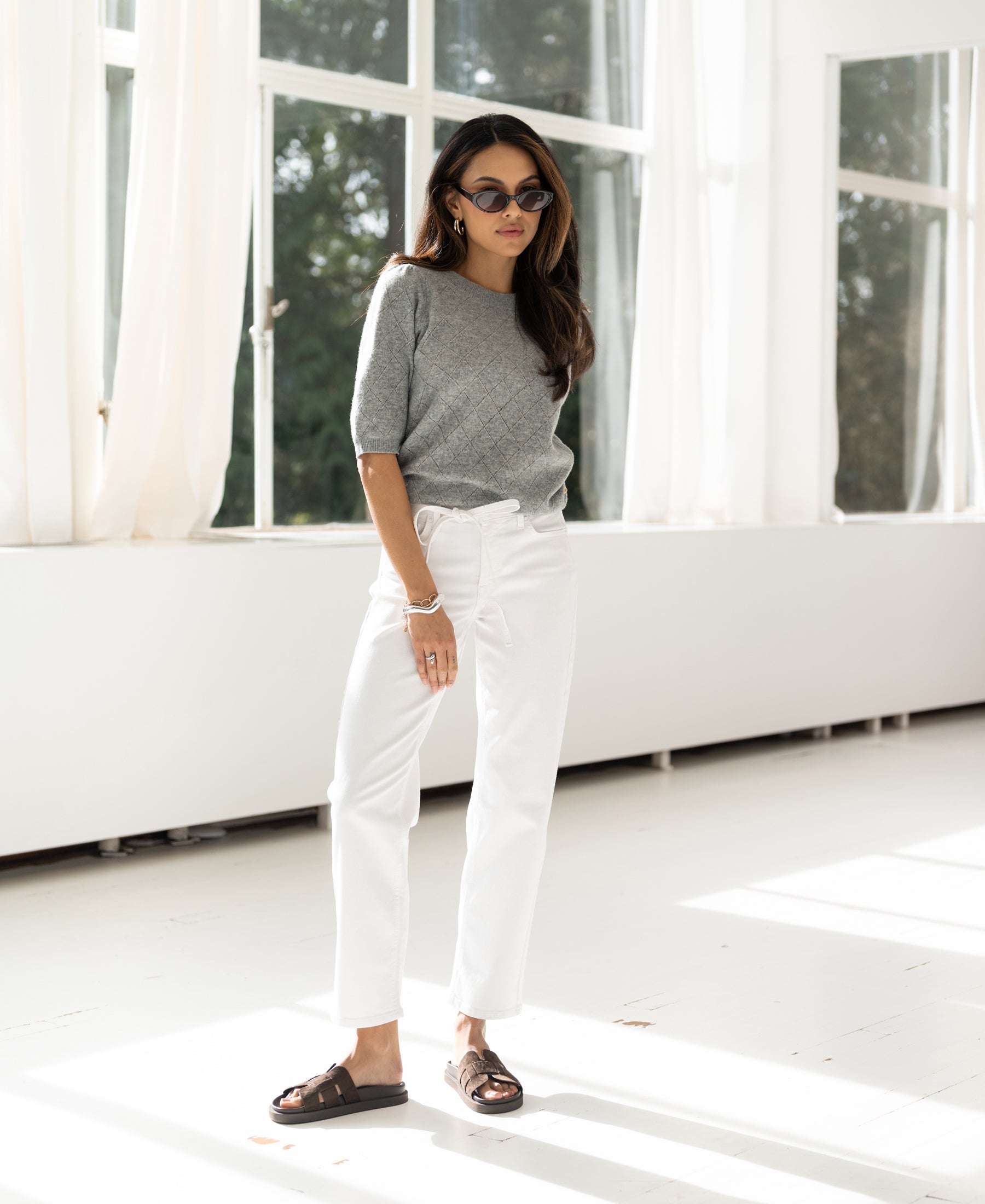 Woman wearing white cropped jeans by Plein Publique stands in a bright room.