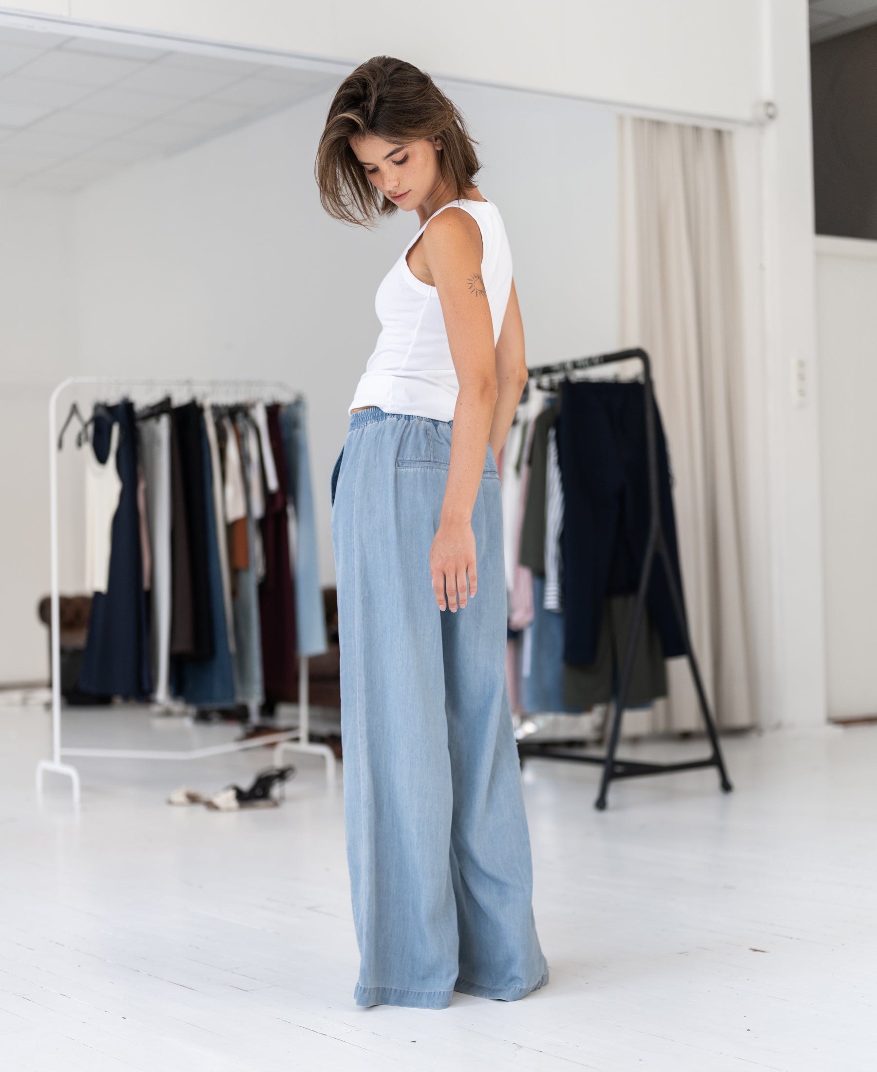 PLEIN PUBLIQUE Tencel jeans LE DEGAS in bleached color, shown worn by a woman standing by clothing racks.
