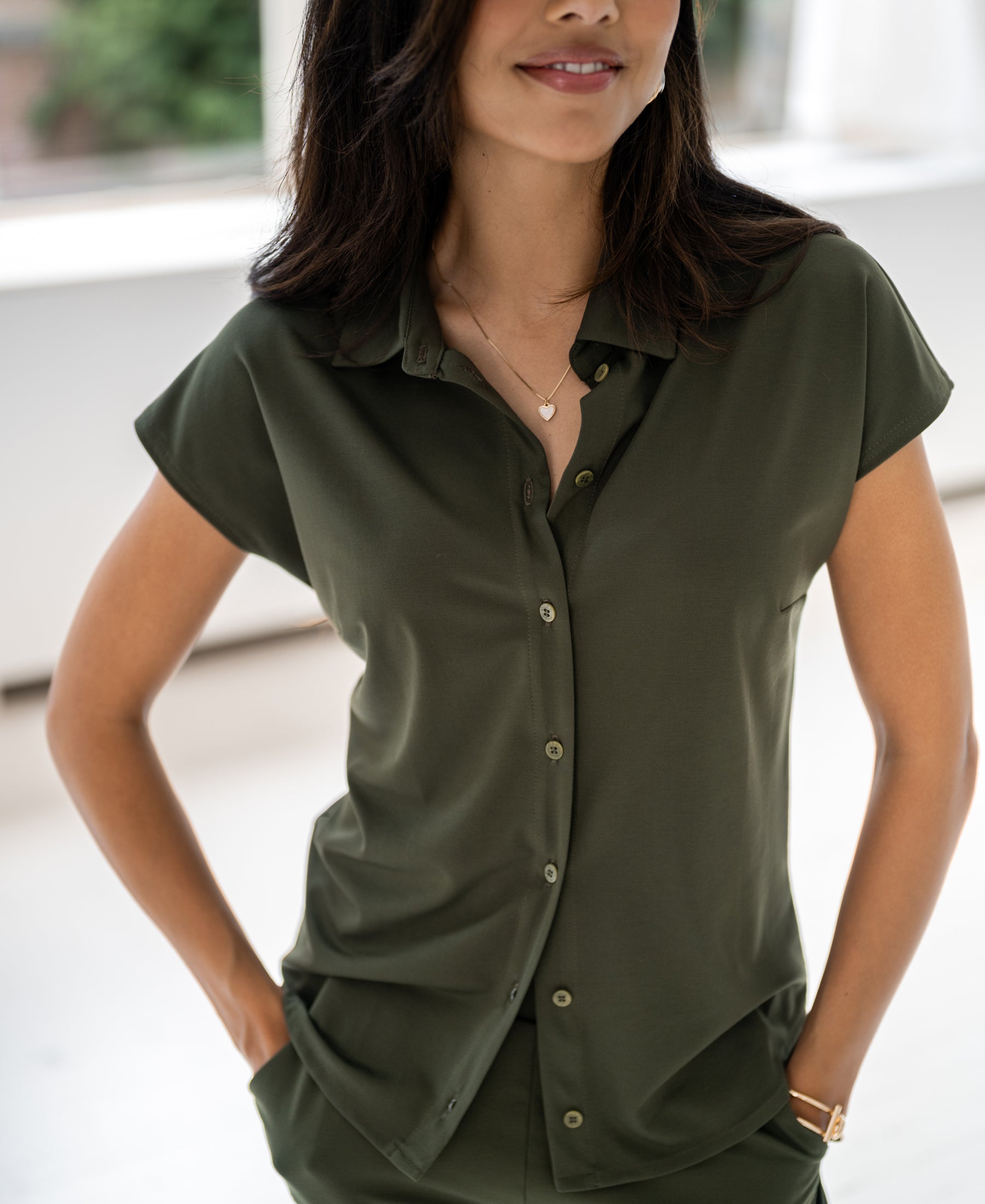 Woman wearing an army green travel blouse, hands in pockets, smiling indoors.