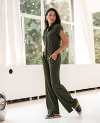 Woman wearing an army green travel blouse stands by large windows, looking to the side with hands in pockets.