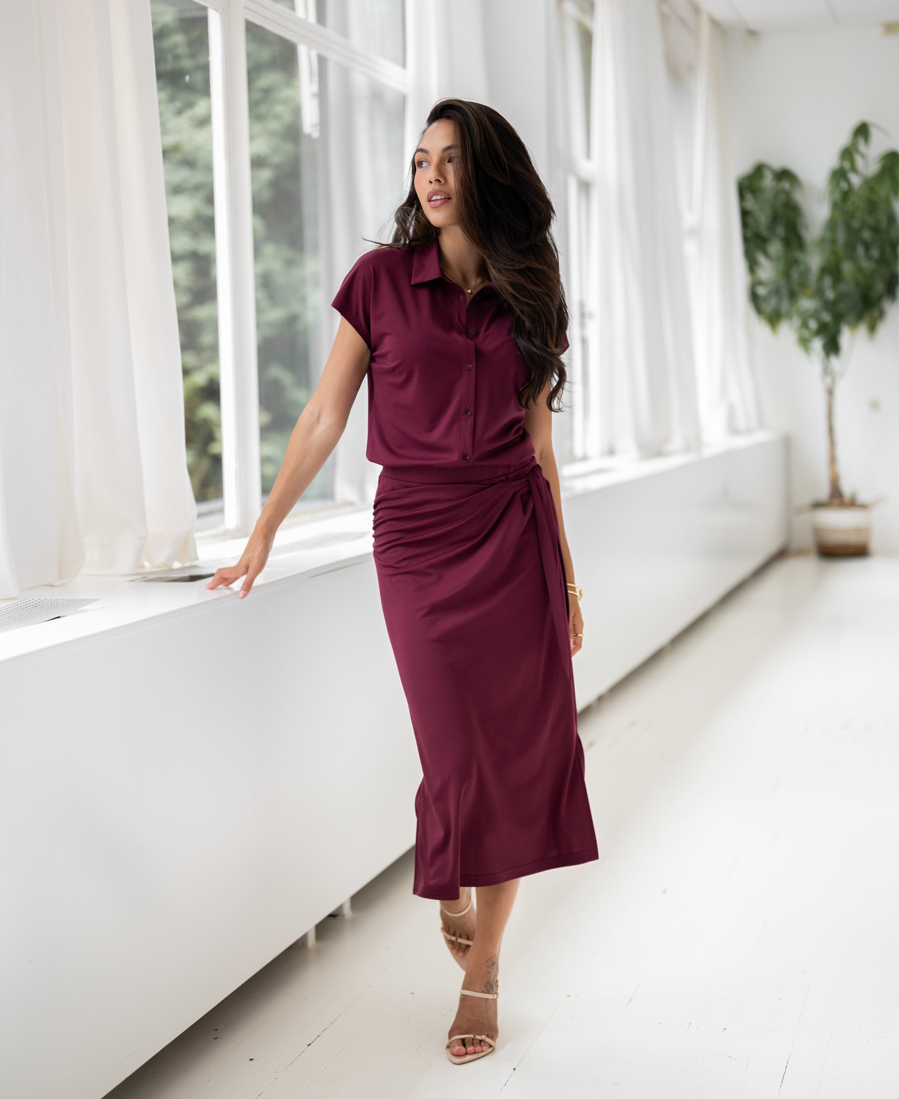 Woman wearing a high-waisted LE MAEL Wine Travel skirt stands by large windows in a bright, plant-filled room.