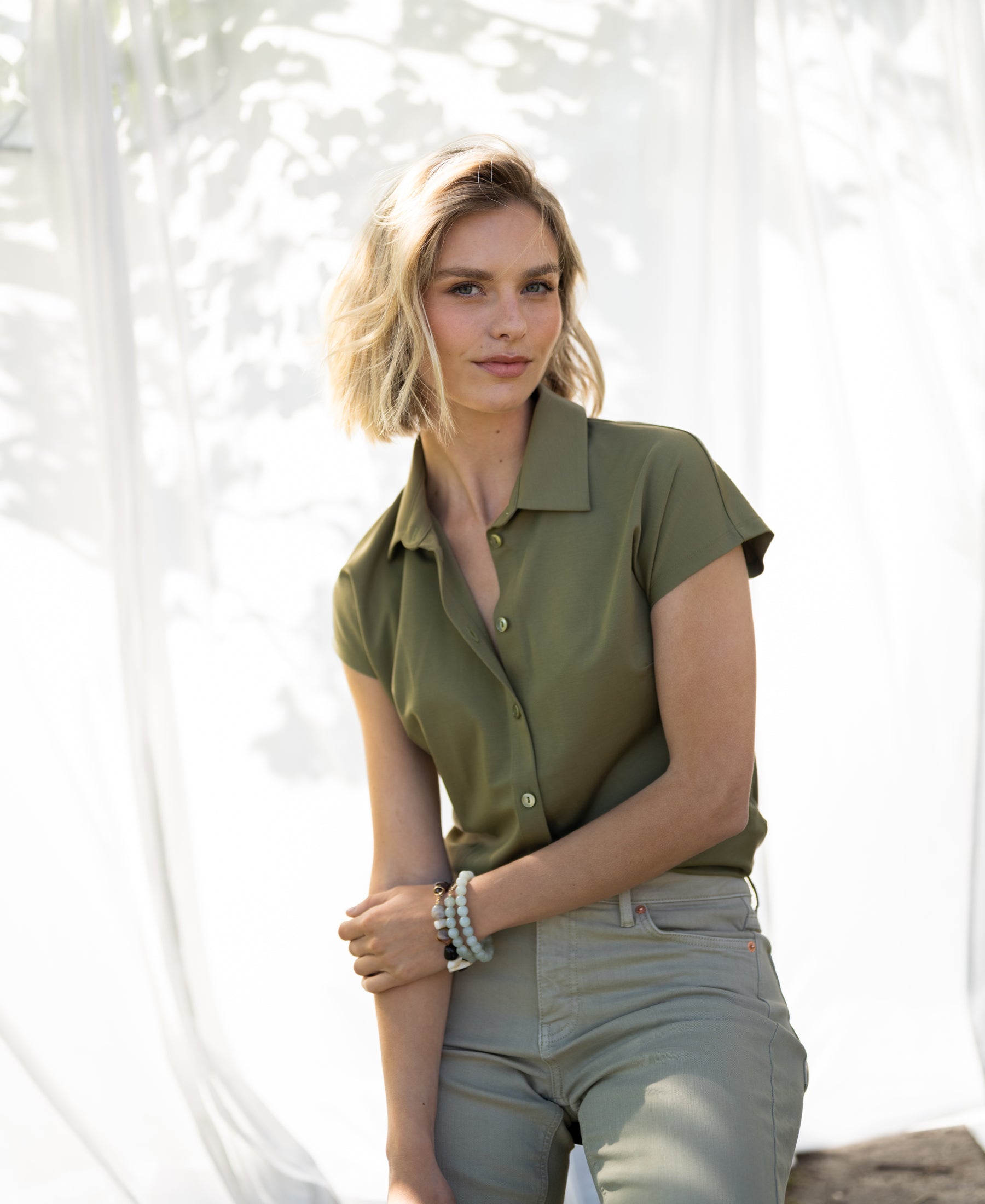 Olive green travel blouse in lightweight fabric, shown in sunlight against a white curtain backdrop.