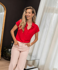 The woman wears the PLEIN PUBLIQUE red LA DELON travel blouse near a window with sheer curtains.