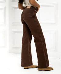 Front view of PLEIN PUBLIQUE brown jeans, shown worn by a woman standing in a white-paneled room.