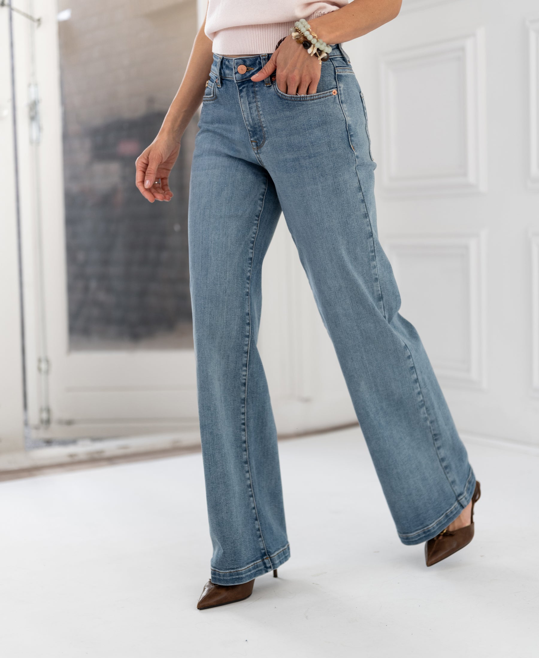 Wide leg jeans in light stone wash by PLEIN PUBLIQUE, shown near white paneled walls and a glass door.