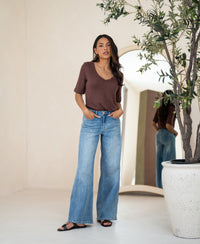 Woman wearing a brown linen T-shirt stands indoors near a potted tree.