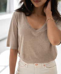 Woman wearing natural-colored linen T-shirt stands indoors, touching her hair and smiling.