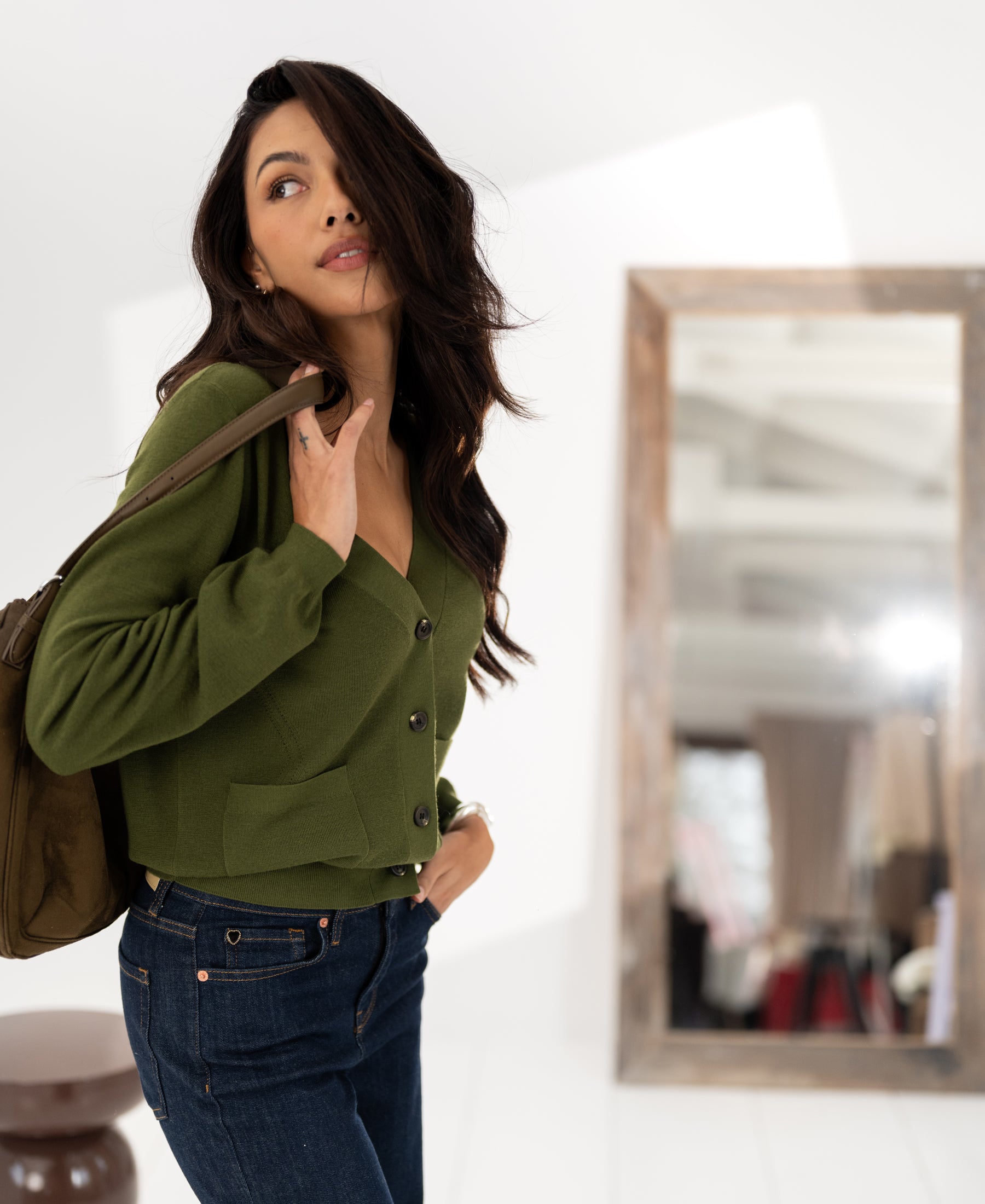 Woman with long dark hair wears the Merino cardigan LA DONNA Olive by PLEIN PUBLIQUE, standing by a mirror.