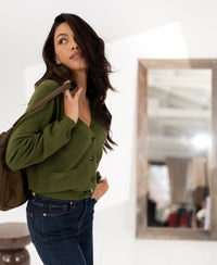 Woman with long dark hair wears the Merino cardigan LA DONNA Olive by PLEIN PUBLIQUE, standing by a mirror.