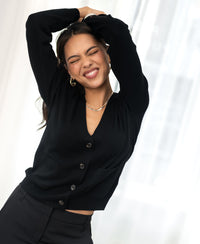 Black merino wool vest, front view, worn by model with eyes closed and arms raised against a bright background.
