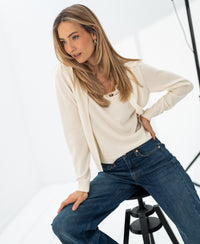 Woman wearing an ivory knit sweater vest sits on a black stool in a bright, minimalist room.