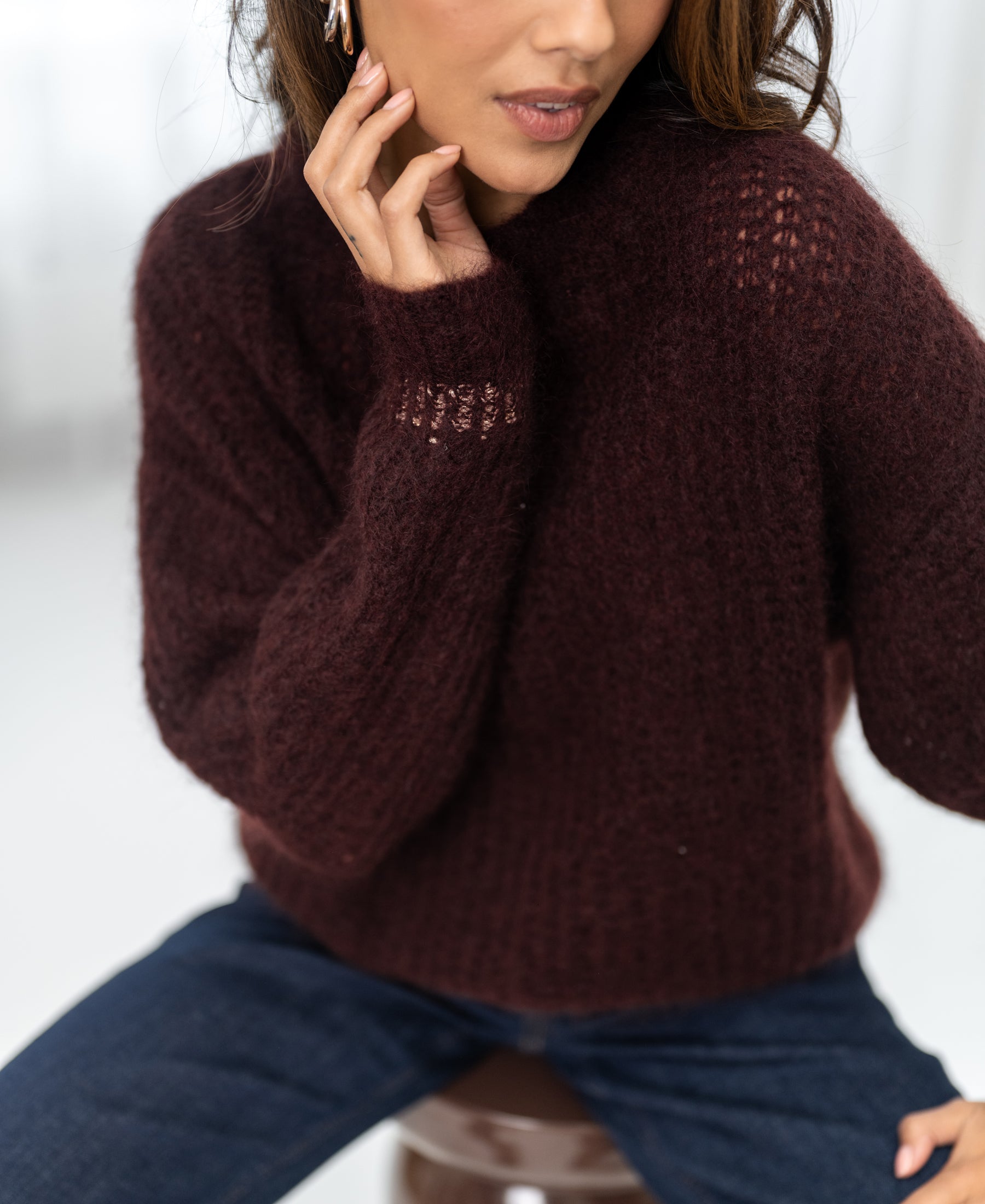 Woman wearing a brownish-red PLEIN PUBLIQUE mohair sweater, one hand gently touching her face.
