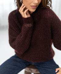 Woman wearing a brownish-red PLEIN PUBLIQUE mohair sweater, one hand gently touching her face.
