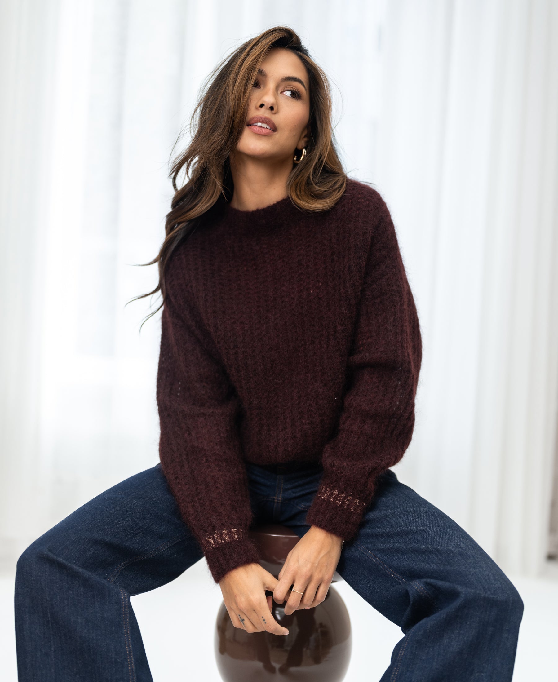 Woman wears the PLEIN PUBLIQUE Mohair sweater LA DOUCE in red-brown, seated on a stool looking upward.