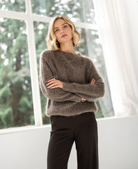 Woman with short blonde hair wears the PLEIN PUBLIQUE mohair sweater LA DOUCE in light brown by a large window.