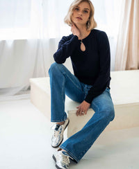 Woman wearing stonewash flared jeans sits indoors, hand on neck, facing camera.