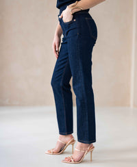Woman wearing cropped jeans in a dark rinsewash stands against a light background.