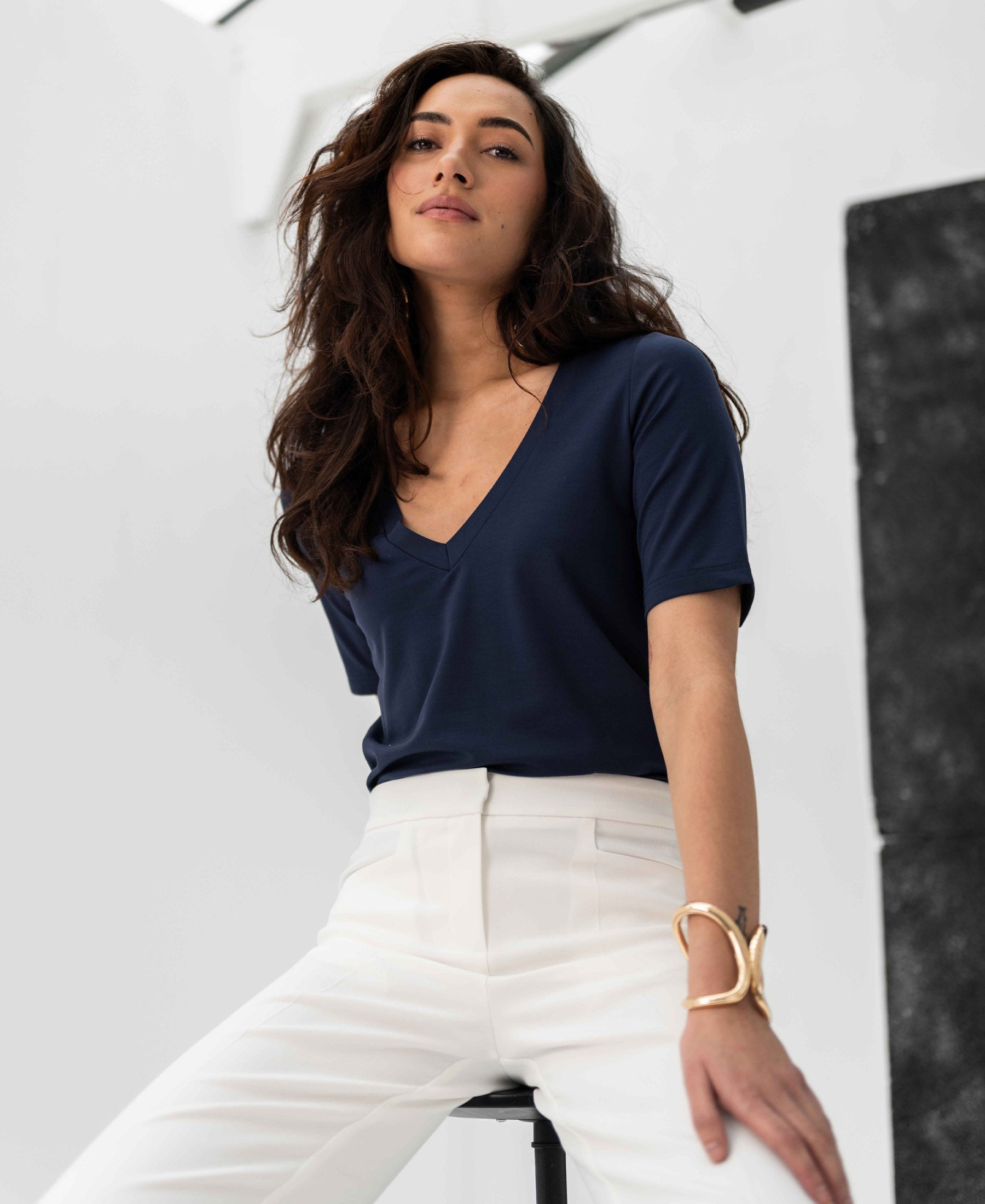 Woman wearing a PLEIN PUBLIQUE LA VERONIQUE V-neck T-shirt in dark blue, seated on a stool.