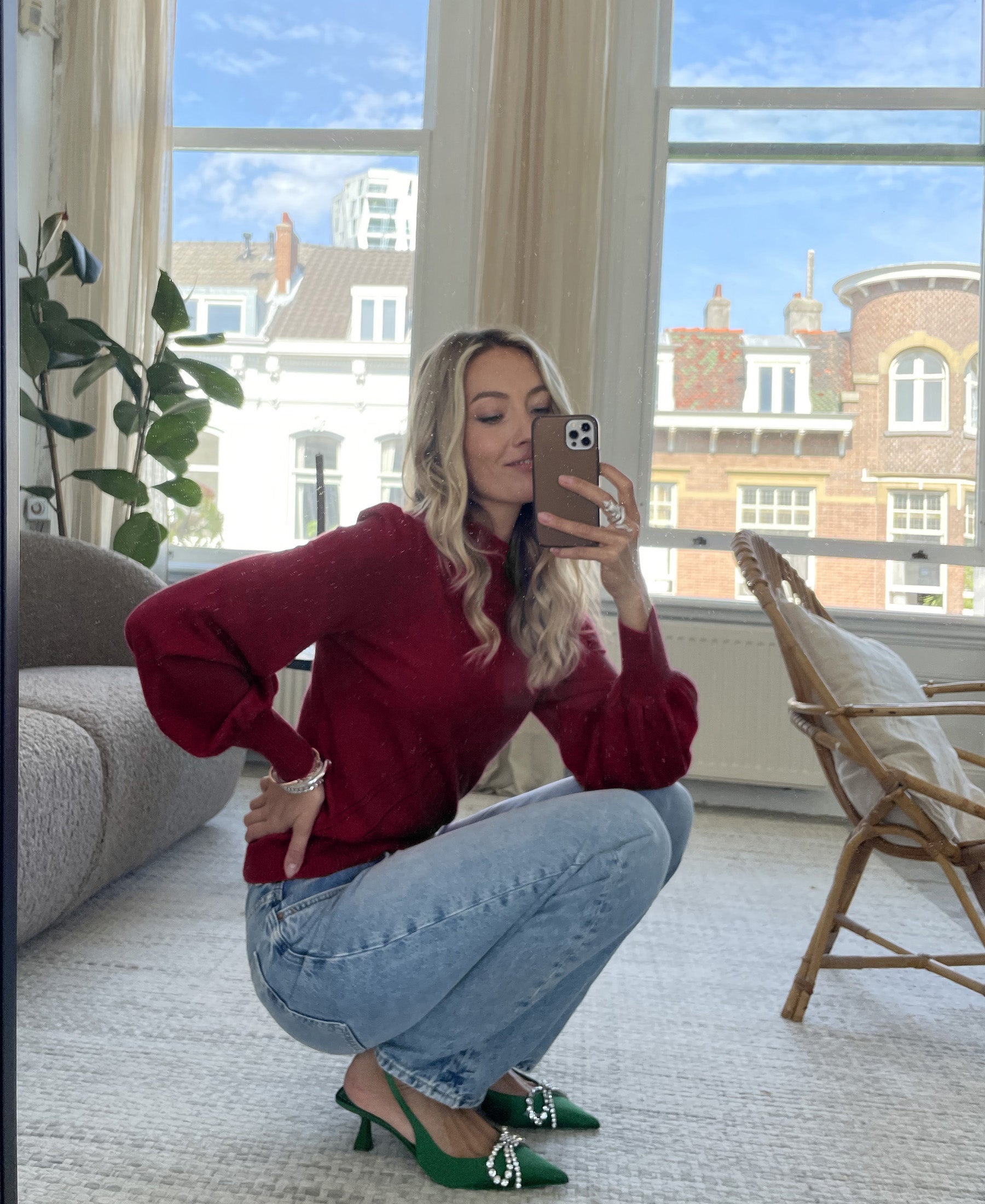 Woman squatting indoors, taking a mirror selfie in the PLEIN PUBLIQUE dark red turtleneck sweater.