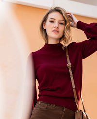 Woman wearing a dark red PLEIN PUBLIQUE LES SALLES turtleneck sweater, standing indoors against a tan wall.