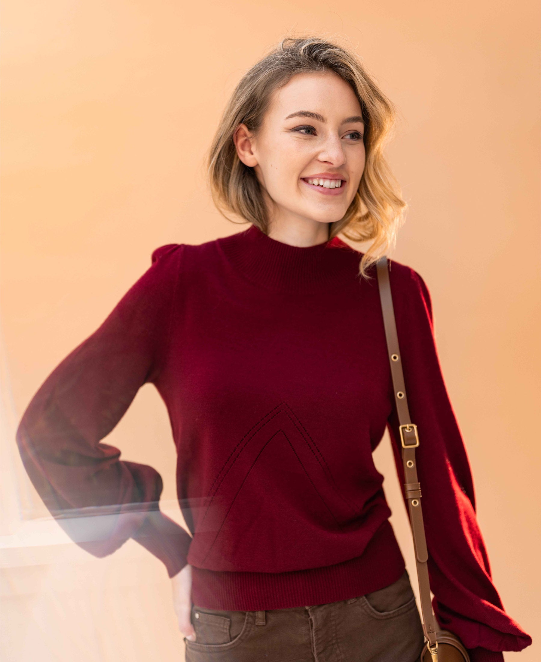 Woman wearing dark red mock neck sweater by a peach wall.