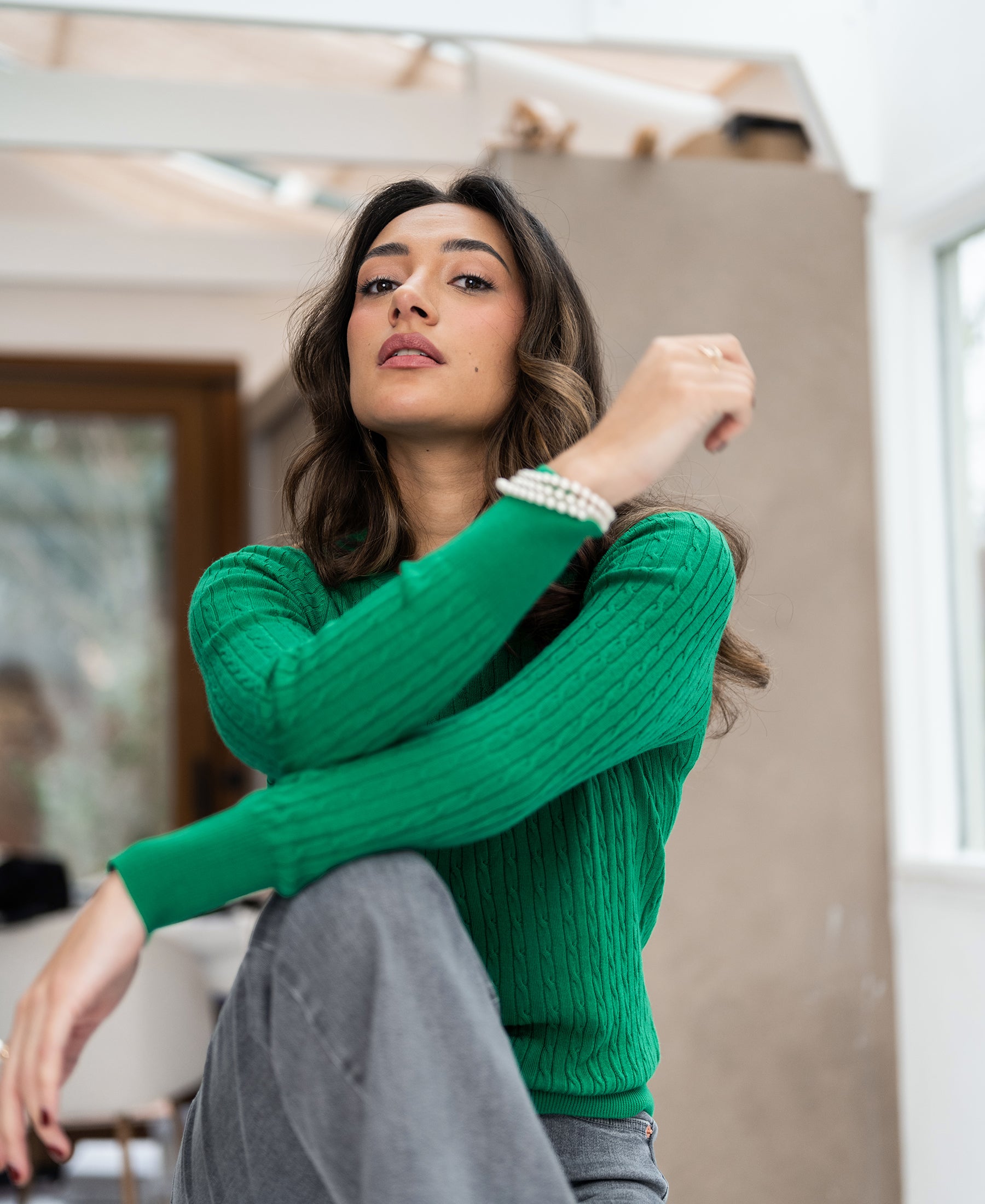 Bright green cable knit sweater shown indoors, front view. Model sits with knee up and arm resting on her leg.