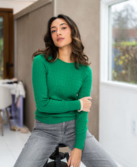 Woman wearing bright green ETHAN cable-knit sweater sitting indoors on a stool.