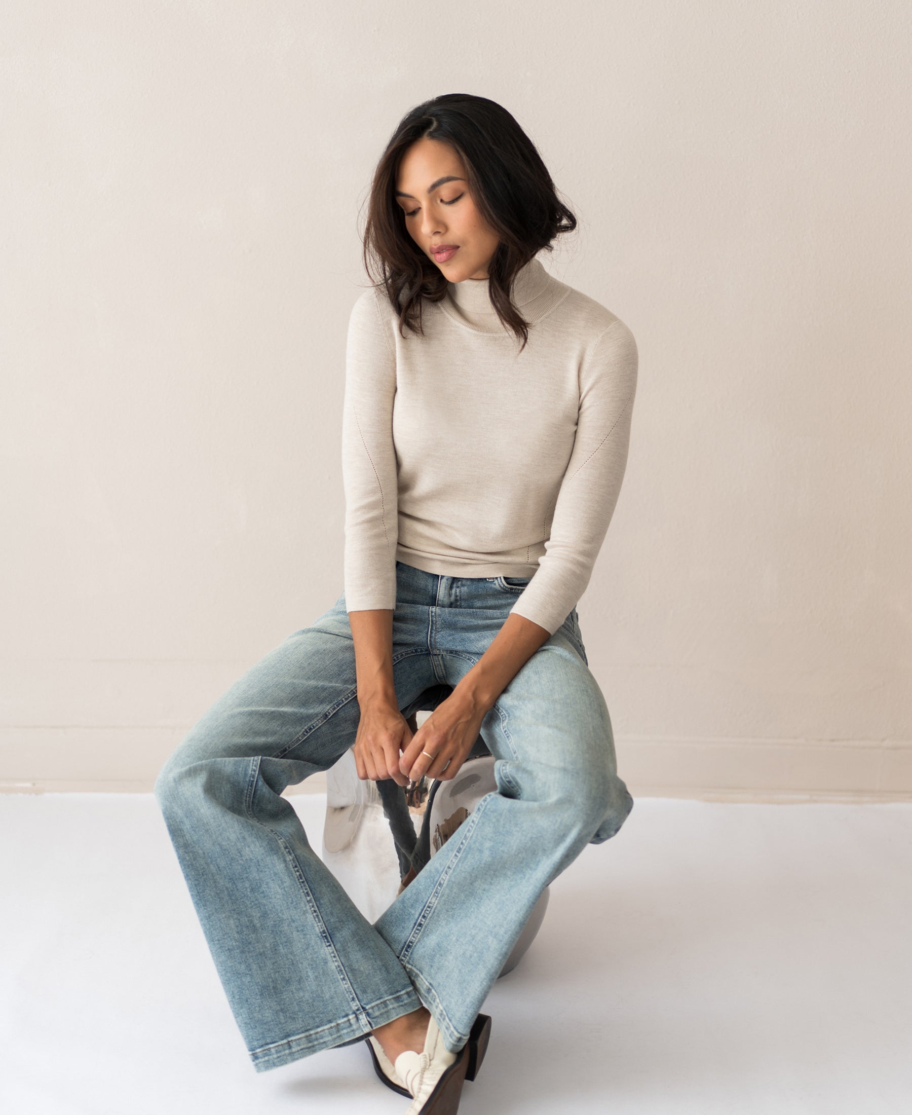 Front view of a woman wearing a beige turtleneck sweater, seated on a stool, looking down with hands clasped.