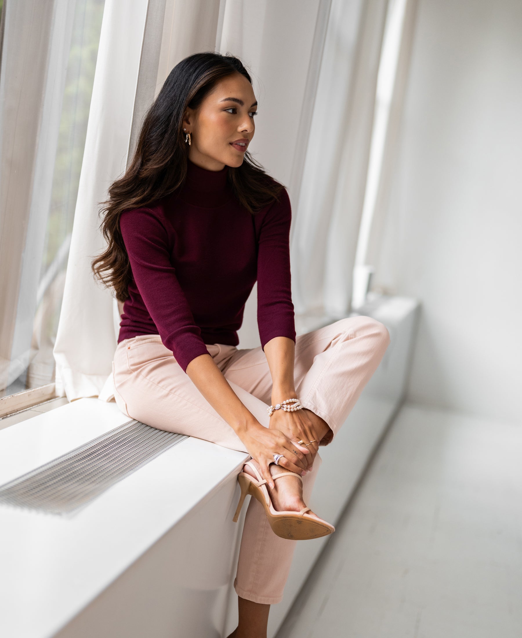 Bordeaux turtleneck sweater by Plein Publique, shown on a model seated on a windowsill.