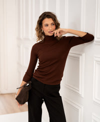 Woman wearing a brown LE FABIAN turtleneck sweater by PLEIN PUBLIQUE stands against a wall.