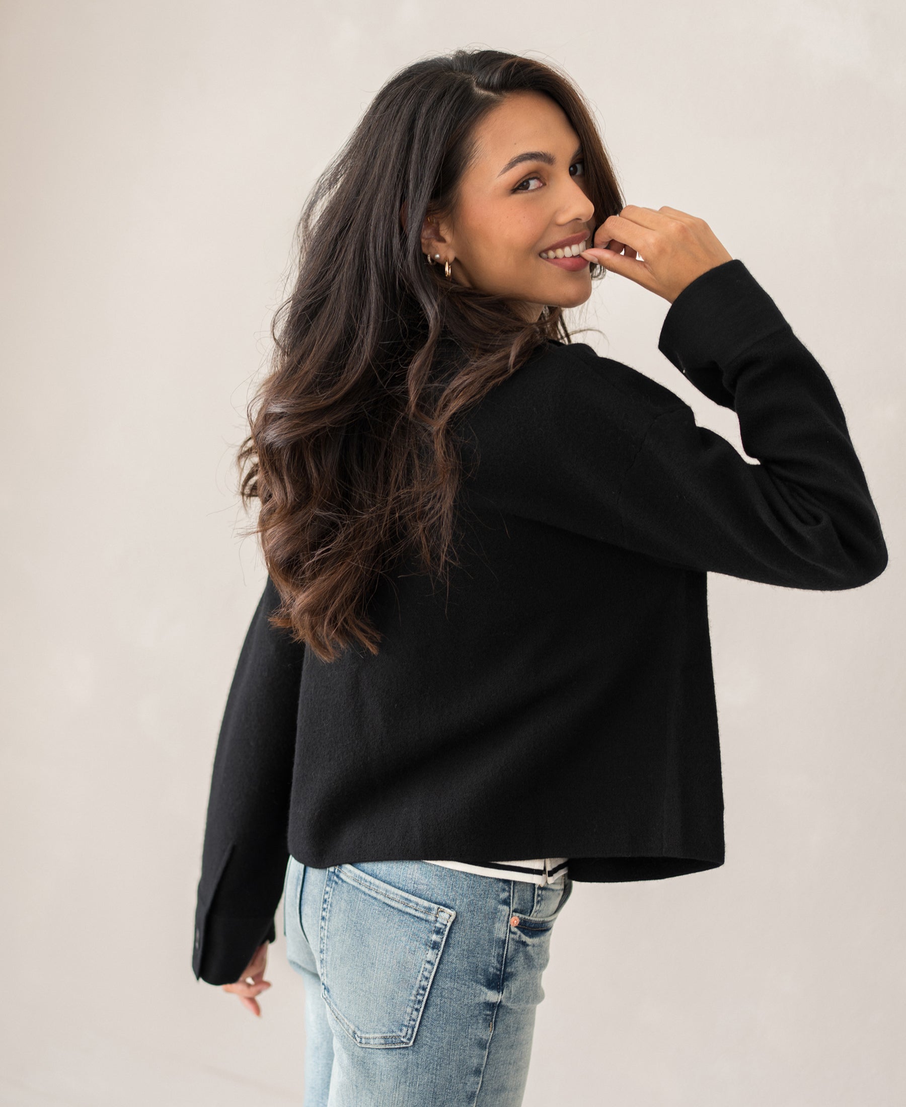 Woman with long dark hair wearing a wool jacket, LA FAYE black, smiling and looking over her shoulder.