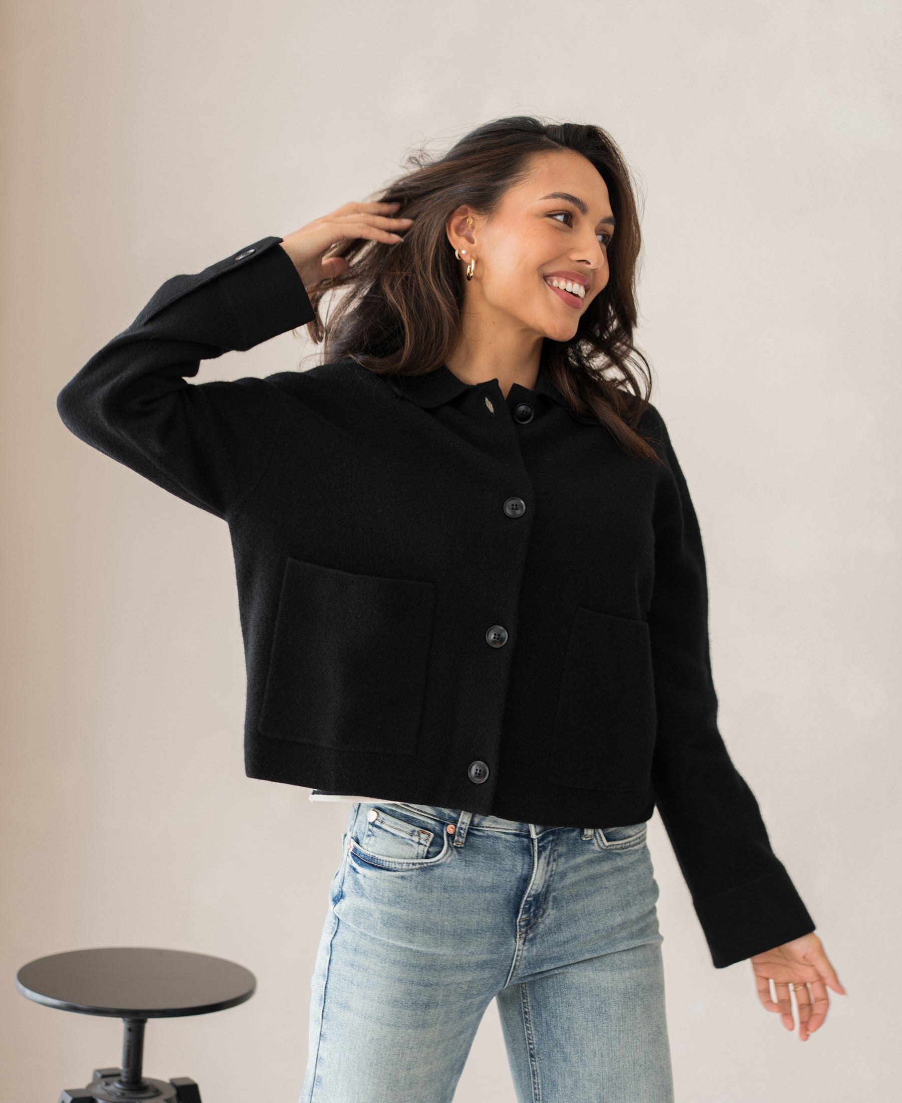 Woman wearing a PLEIN PUBLIQUE wool jacket LA FAYE black, indoors with a stool nearby.