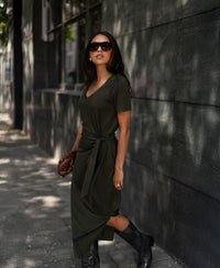 Woman wears a dark olive LA FILICIA long dress by PLEIN PUBLIQUE, walking by a dark wall.