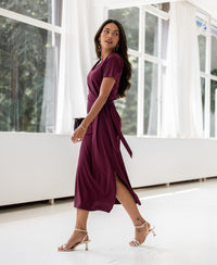 Woman wearing a long wine-colored dress called The Filicia walking indoors with large windows and white curtains.