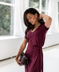 Woman wearing a long wine-colored dress stands indoors as sunlight streams in.