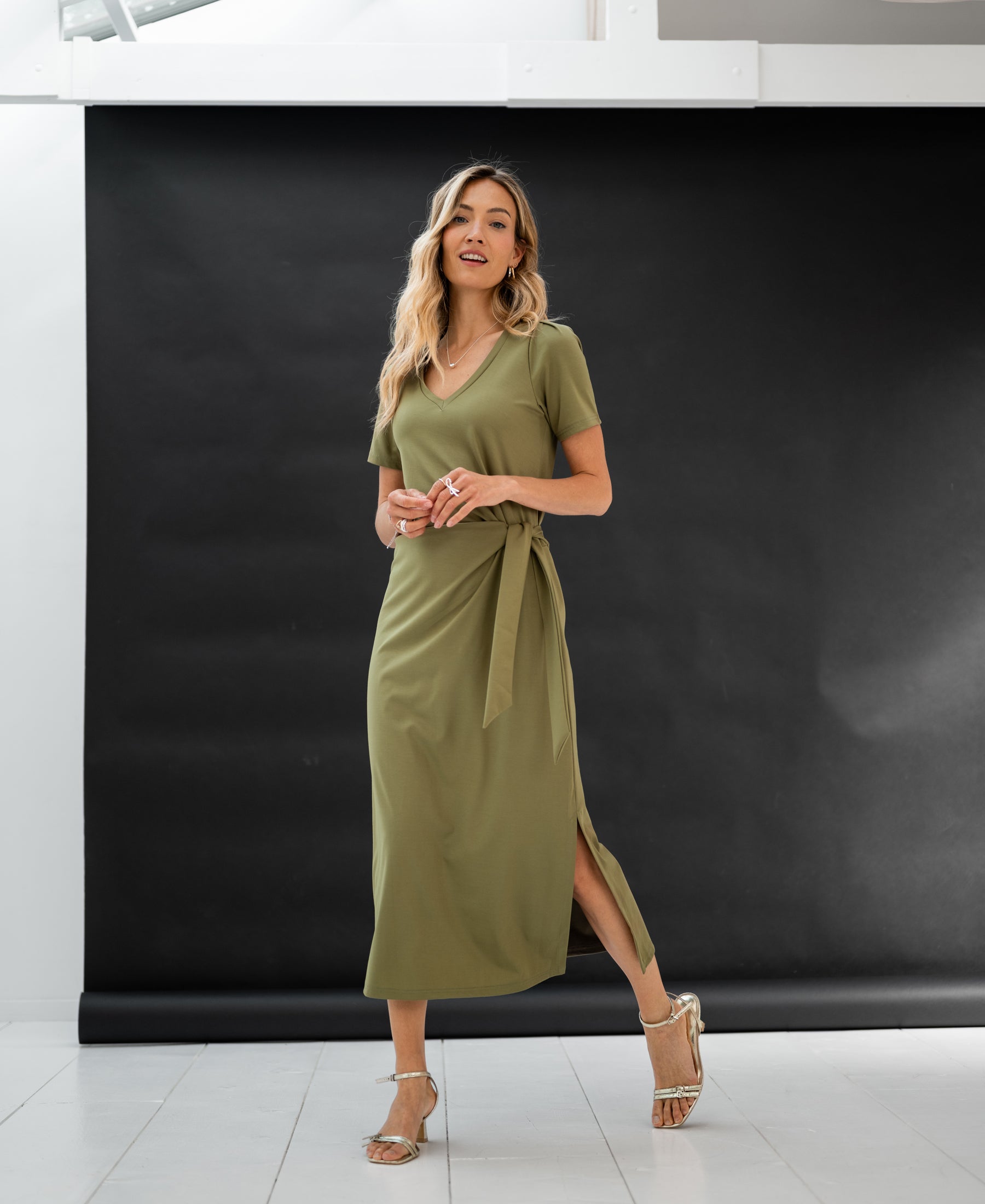 Woman wears the long dress LA FILICIA olive green by PLEIN PUBLIQUE in a bright studio with a black backdrop.