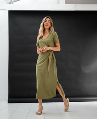 Woman wears the long dress LA FILICIA olive green by PLEIN PUBLIQUE in a bright studio with a black backdrop.