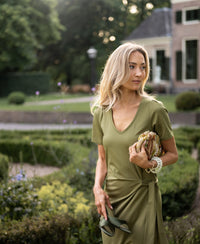 Woman wearing the LA FILICIA olive green long dress by PLEIN PUBLIQUE stands in a sunlit garden.