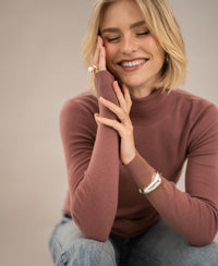 Woman wearing a PLEIN PUBLIQUE Merino wool turtleneck sweater in dusty pink, hand resting on her cheek.