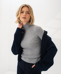 A woman wears a PLEIN PUBLIQUE Merino wool turtleneck sweater in Feathergrey, shown against a minimal backdrop.