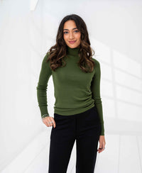 Woman wearing an olive green PLEIN PUBLIQUE merino wool turtleneck sweater, standing and smiling in a bright room.