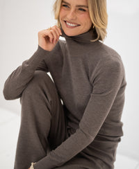 Woman wearing a dark sand-colored merino turtleneck sweater, smiling with hand resting on her chin.
