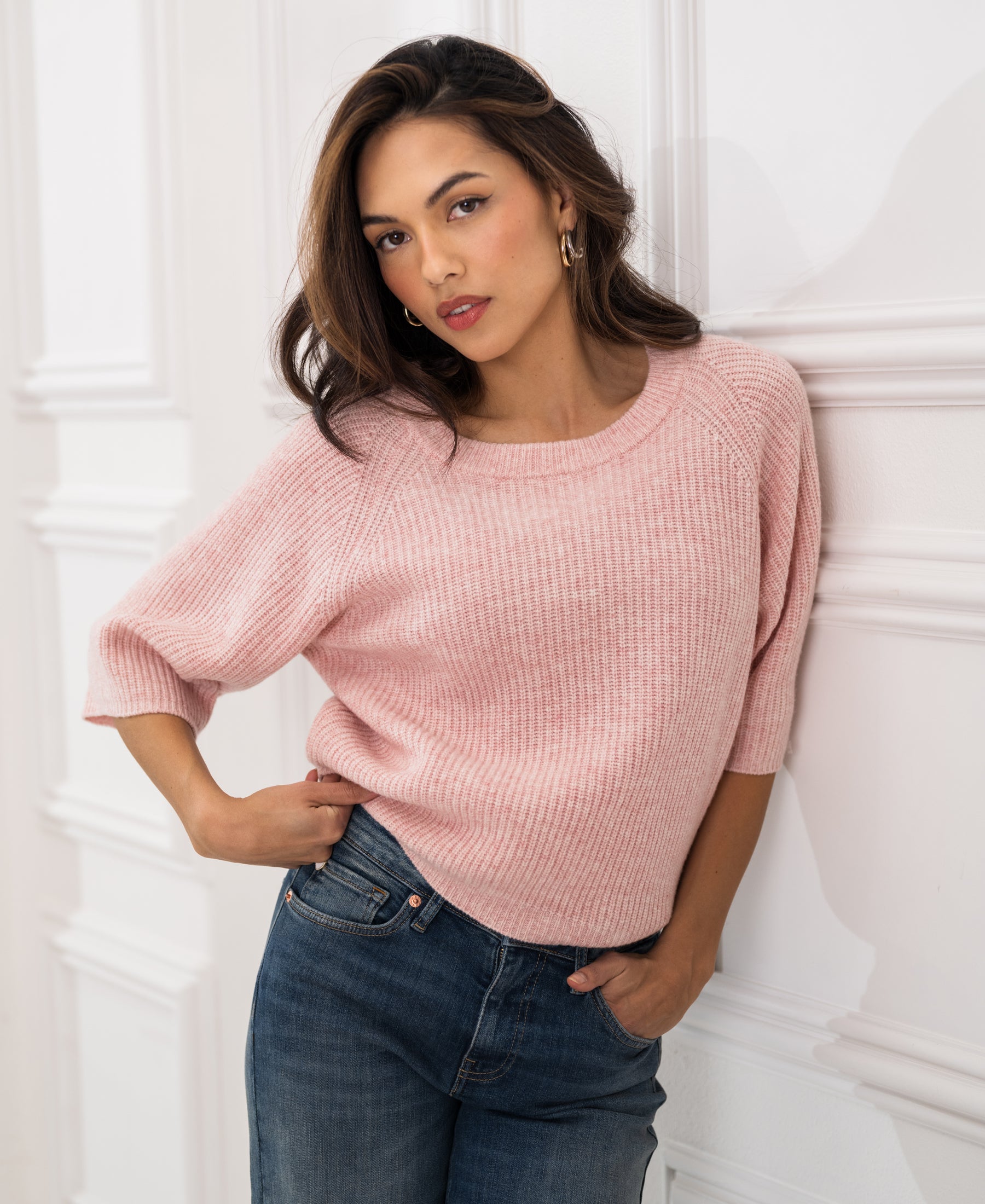 Woman wearing a pink PLEIN PUBLIQUE sweater stands against a white wall, one hand in her pocket.