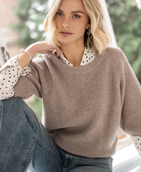 Warm taupe sweater by PLEIN PUBLIQUE, Trui LA FRAMBOISE, shown indoors. Green trees visible outside the window.