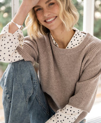 Warm taupe LA FRAMBOISE sweater by PLEIN PUBLIQUE, shown on a model seated by a window with hand on head.