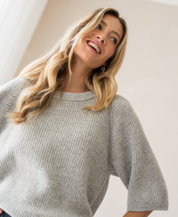 Light grey knit sweater, shown on model standing in a softly lit room.