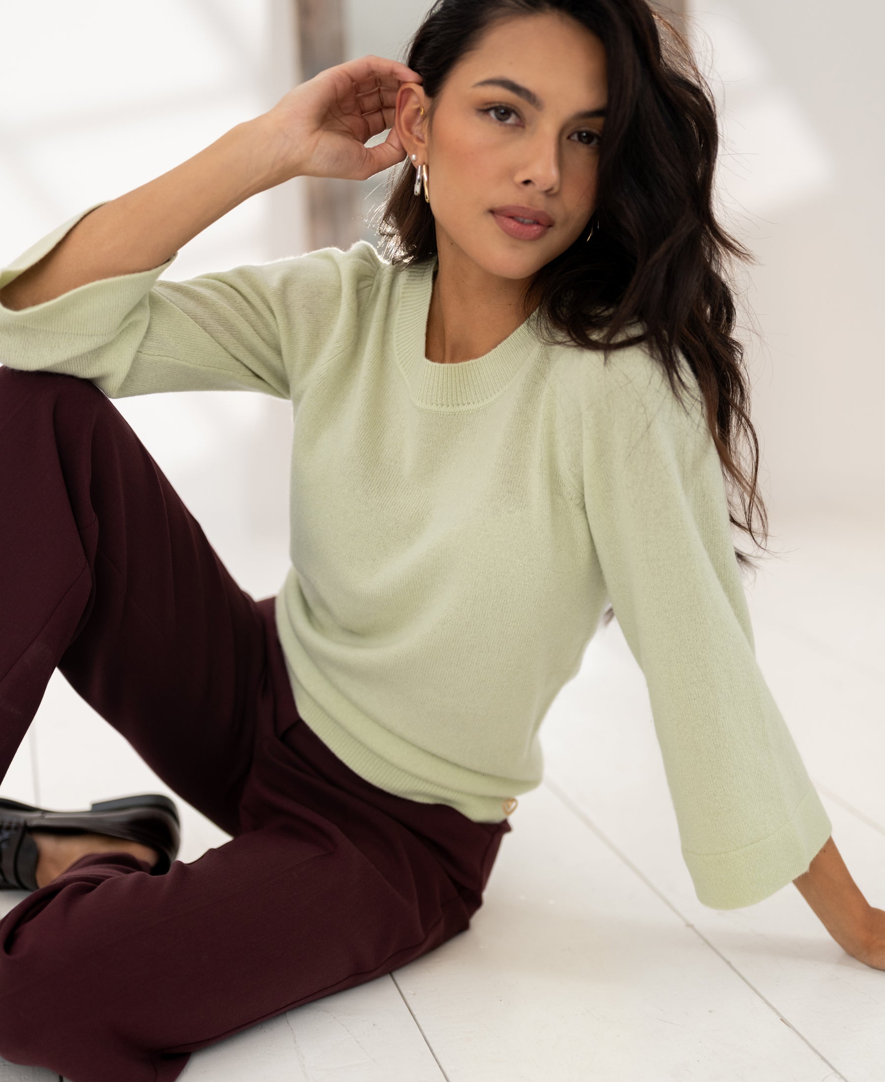 Light green merino wool sweater by PLEIN PUBLIQUE, shown worn by a woman seated on a white floor, facing the camera.